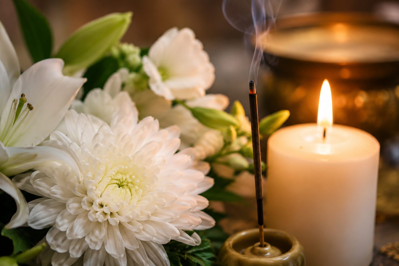 Close-up of white chrysanthemums and lilies beside a lit candle and a stick of incense, symbolizing shared traditions of remembrance